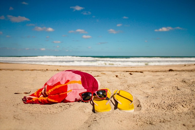 chinelo, toalha e óculos de sol na areia da praia, com mar e céu azul ao fundo