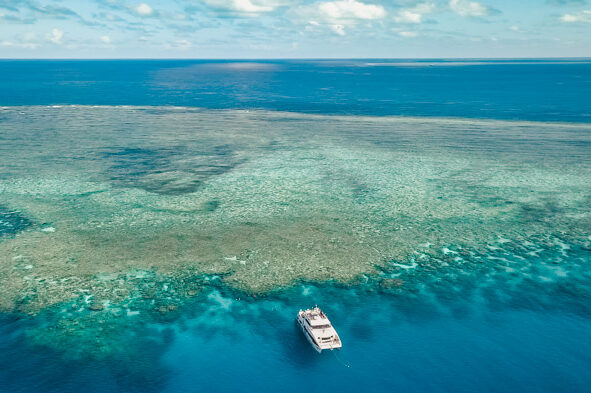 Imagem aérea da Grande Barreira de Corais, barco parado na frente da barreira, mar azul.
