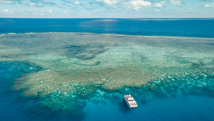 Imagem aérea da Grande Barreira de Corais, barco parado na frente da barreira, mar azul.