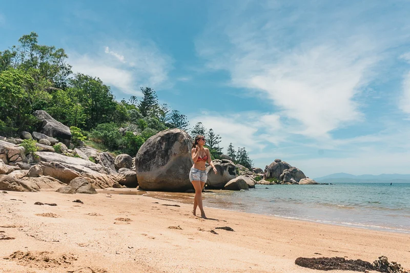 Tatá caminhando pela praia de Picnic Bay, em Magnetic Island, com mar azul e vegetação ao fundo.