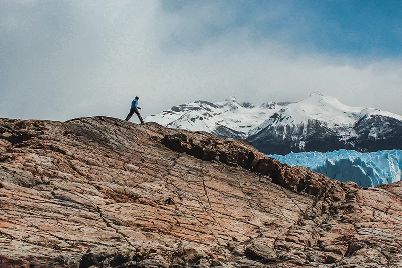 Viajante caminhando por uma formação rochosa diante de montanhas nevadas na Patagônia.