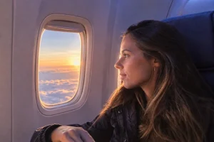 Tatá sentada na janela do avião, observando o céu durante o voo ao pôr do sol.