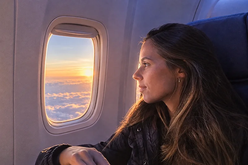 Tatá sentada na janela do avião, observando o céu durante o voo ao pôr do sol.