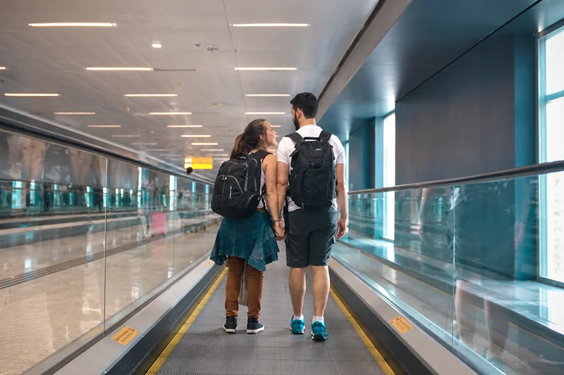 Casal caminhando junto por um corredor de aeroporto com mochilas durante uma viagem.